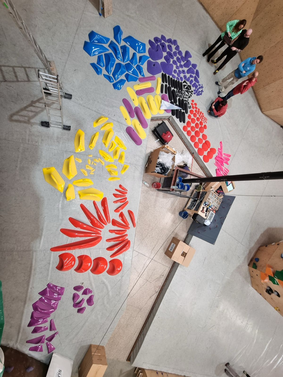 colorful climbing holds on the floor. A few people on the right side.